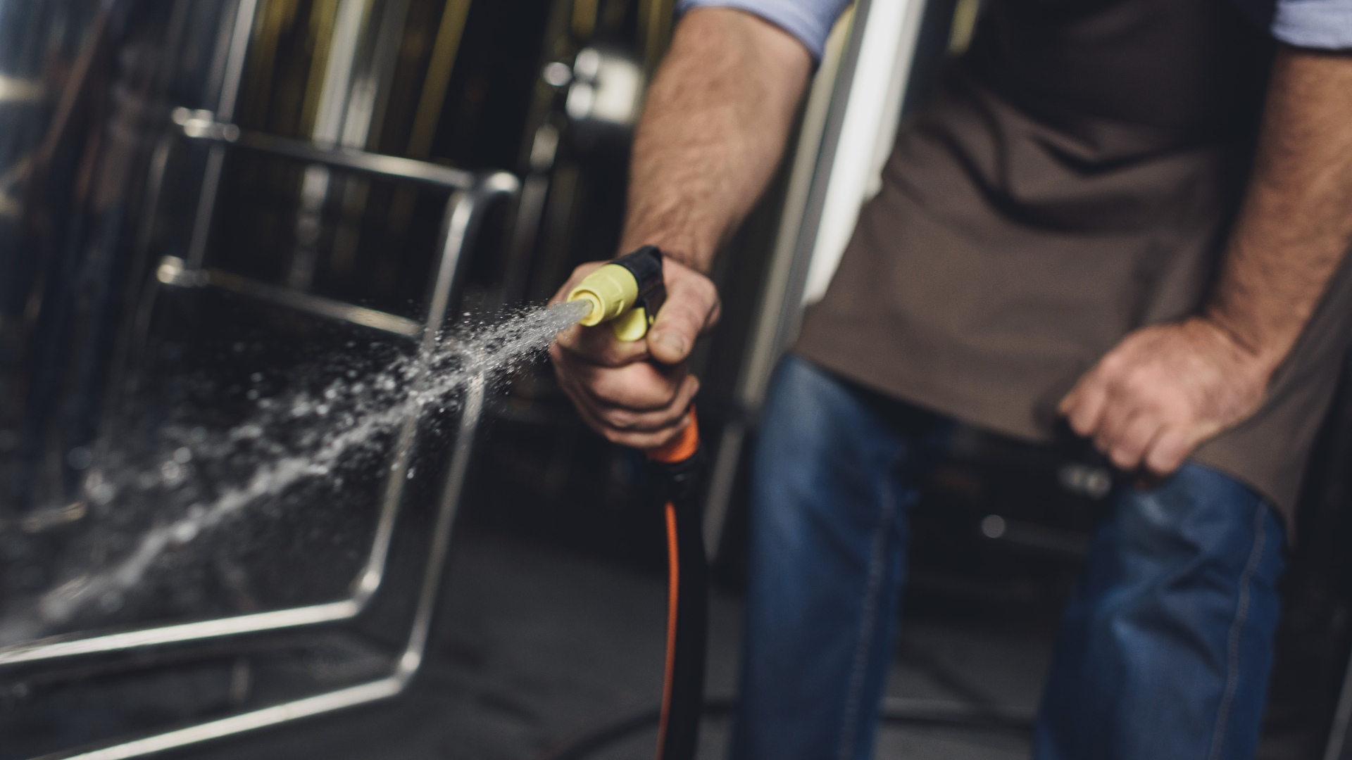 A man spraying brewery equipment with a cleaner from a hose.